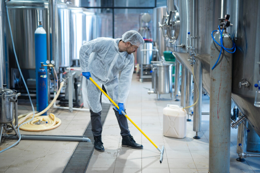 Person in Schutzkleidung reinigt den Boden in einer Produktionsanlage mit Edelstahlbehältern.